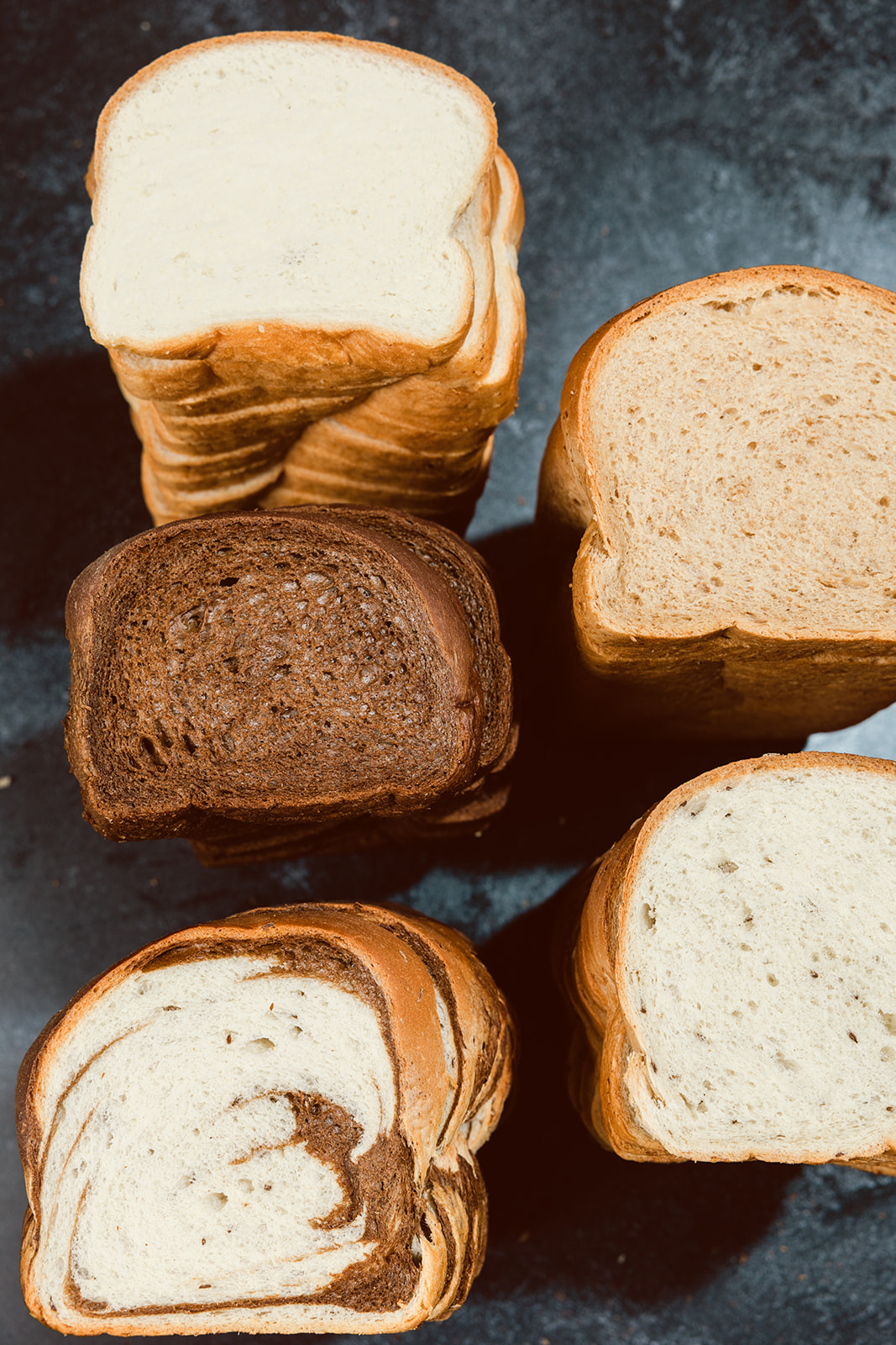 Top-down view of sliced La Segunda basket bread varieties — white, wheat, rye, marble, and sourdough loaves cross-sectioned on a dark surface.