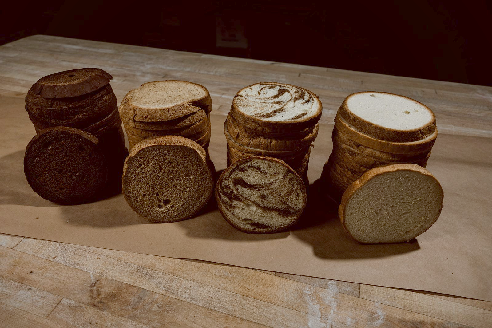 Cross-section slices of pumpernickel, rye, marble, and sourdough deli loaves arranged on a wooden bench against a dark background.