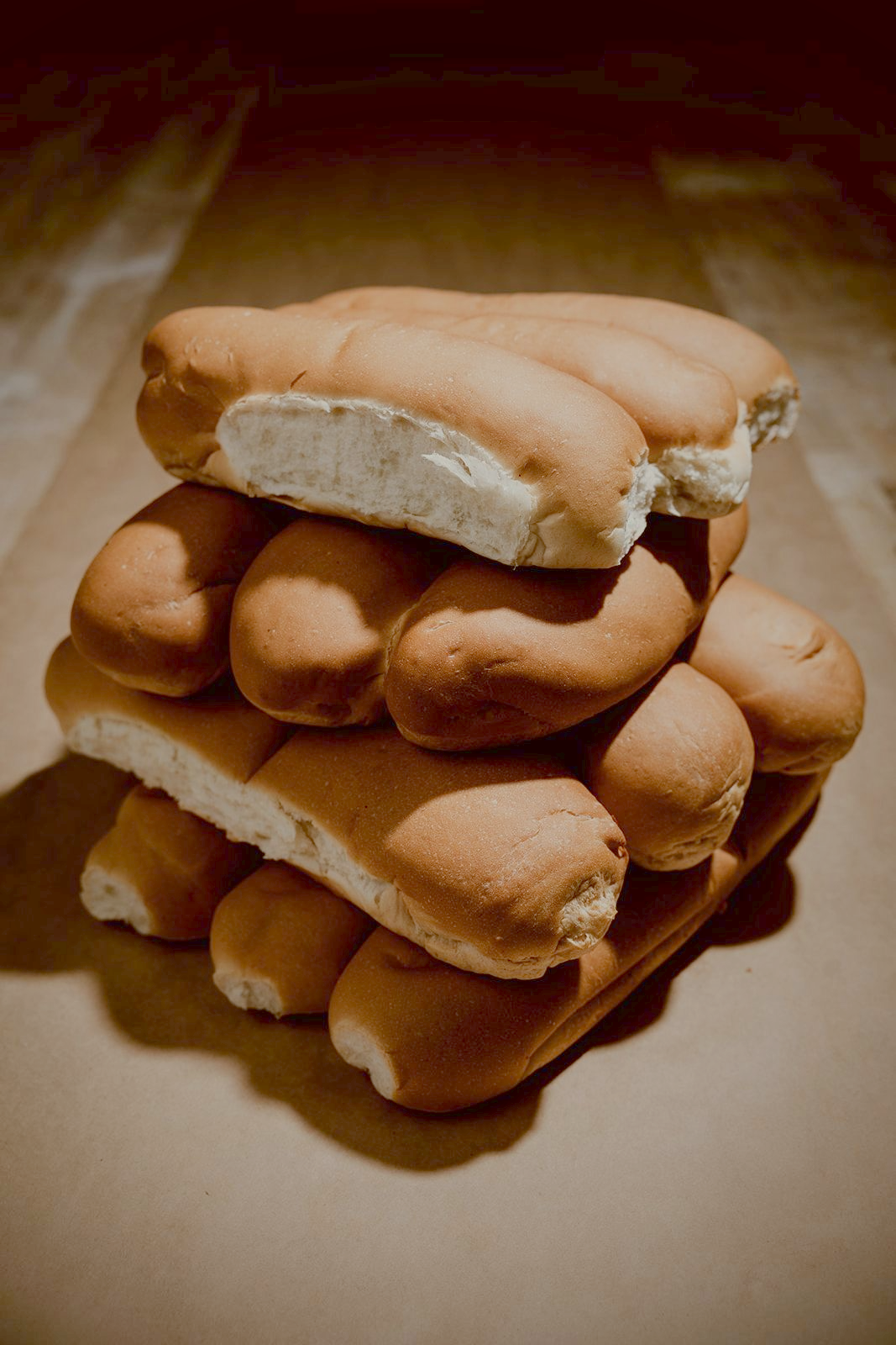 A stack of fresh 8-inch and 12-inch hoagie rolls in soft warm bakery light against a deep shadow background.