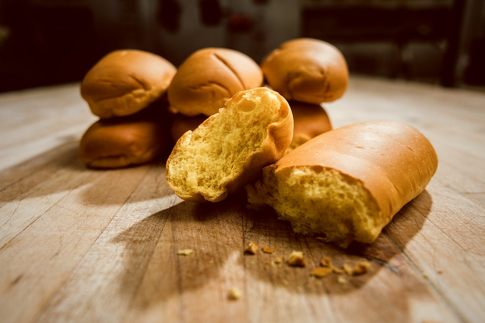 A cluster of golden Media Noche hoagie rolls with glossy eggwash crust, lit by warm low light against a deep shadow background.